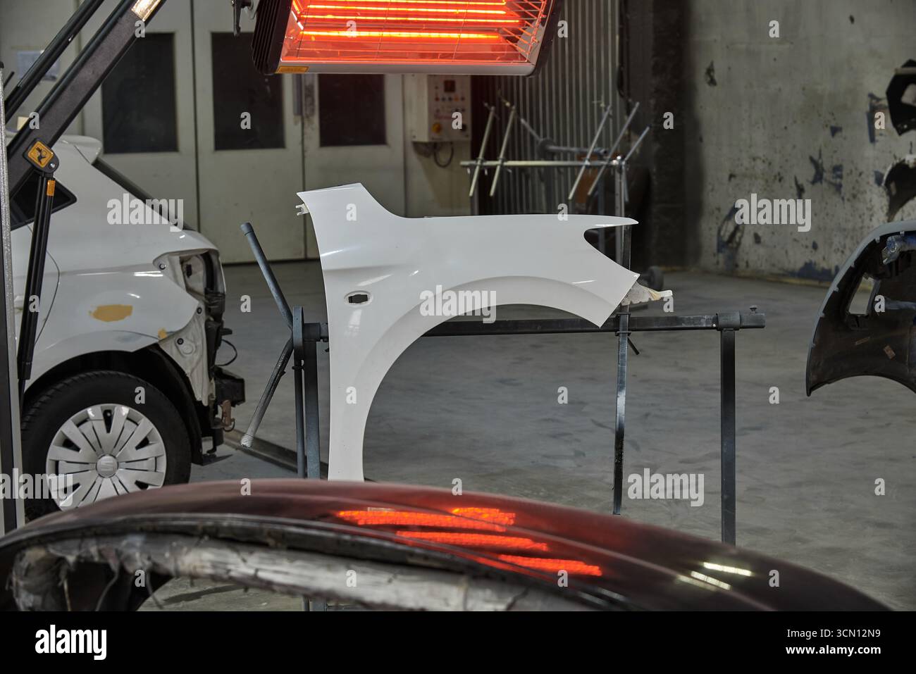 Ein weißer Autokotflügel unter einer roten Wärmelampe in einer Werkstatt, mit Werkzeugen, Lackspritzern und einem beschädigten Fahrzeug in der Nähe, das eine aktive Karosserie zeigt Stockfoto