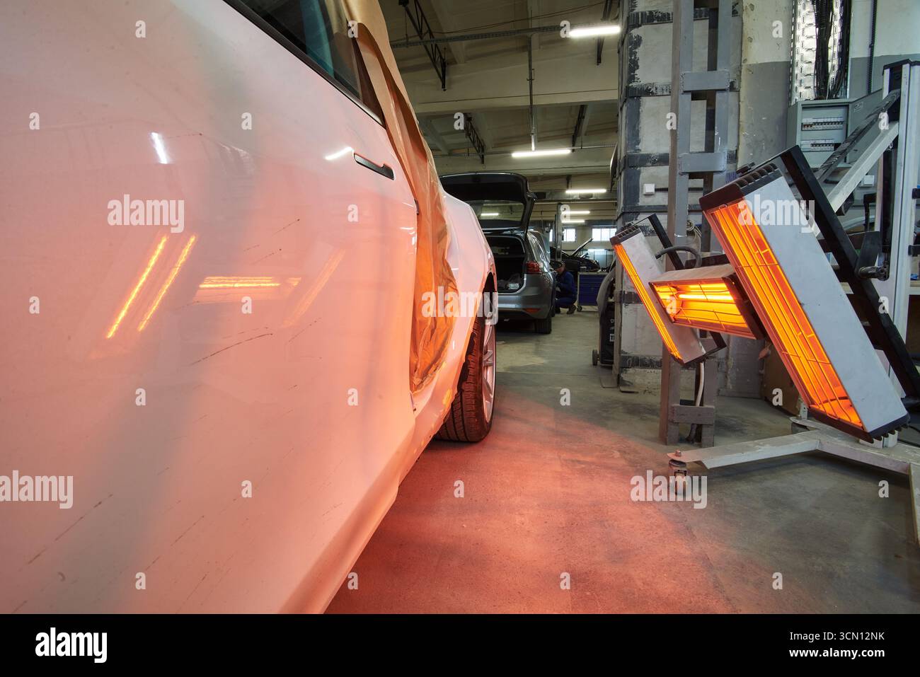 In einer Werkstatt steht ein weißes Auto mit seitlichen Schäden, beleuchtet von roten Infrarot-Wärmeleuchten zum Aushärten von Farbe, umgeben von Werkzeugen und anderen Fahrzeugen Stockfoto