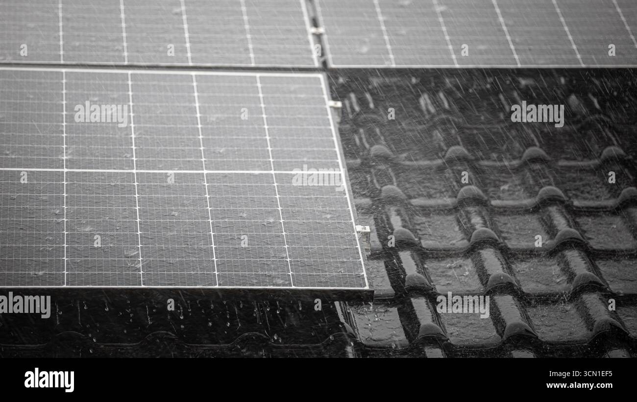 Schwarzes Solarpanel auf glänzend schwarzen Dachziegeln bei starkem Regenguss, intensiver Regen auf der Platte, Teilansicht des dahinter liegenden Daches, Format 16:9 Stockfoto