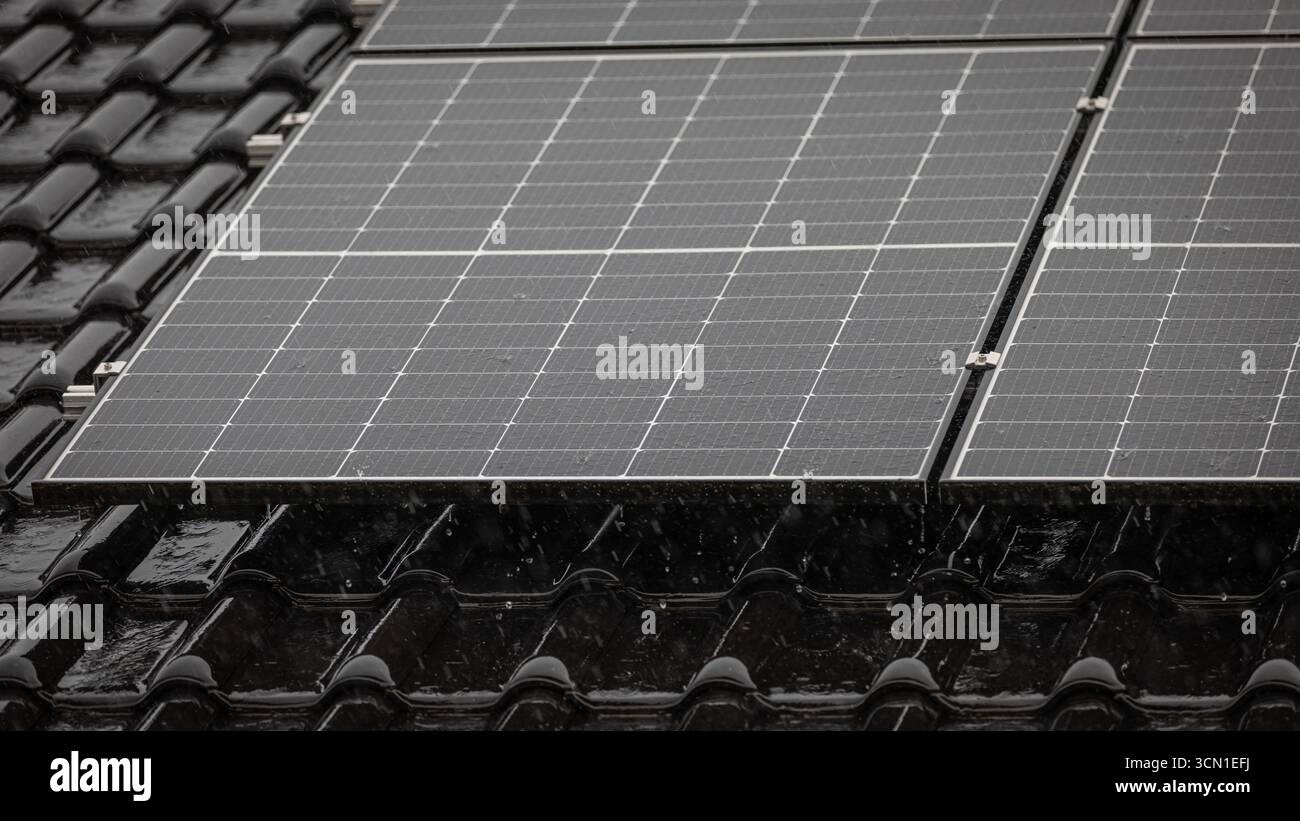 Schwarzes Solarpanel auf glänzend schwarzen Dachziegeln bei starkem Regen, tropfenförmige Tropfen auf der Platte, Teilansicht des dahinter liegenden Daches, Format 16:9 Stockfoto