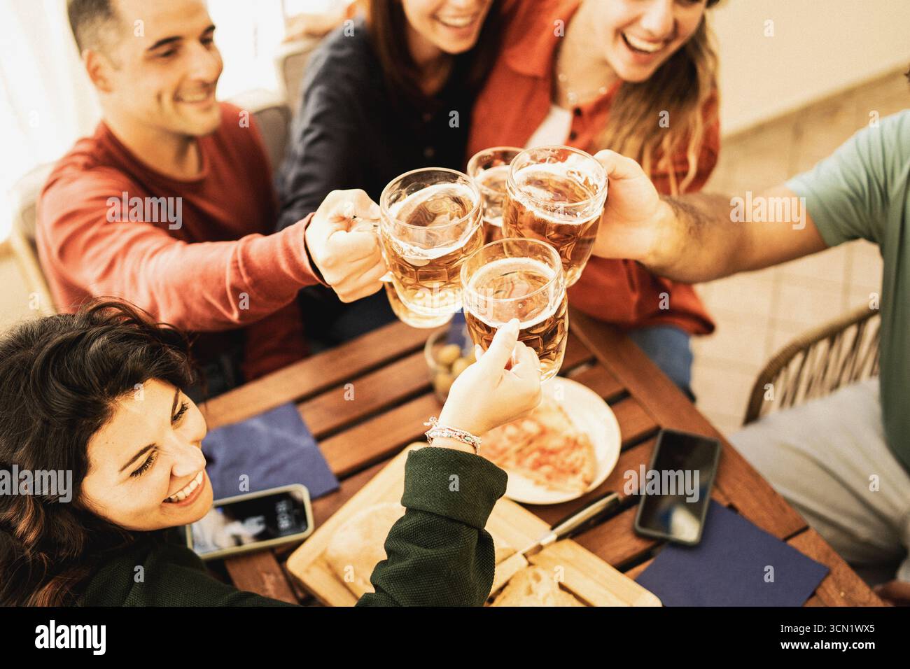 Aus einem hohen Winkel stoßen eine Gruppe glücklicher Freunde ihre Bierbecher in einem Toast über einem Holztisch mit Pizza an, um einen Moment der Zweisamkeit zu feiern. Stockfoto