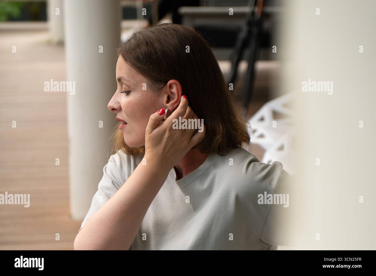 Eine Frau in einem hellgrauen T-Shirt passt ihr kurzes Haar an, zeigt silberne Creolen und rote Maniküre auf einer Terrasse Stockfoto
