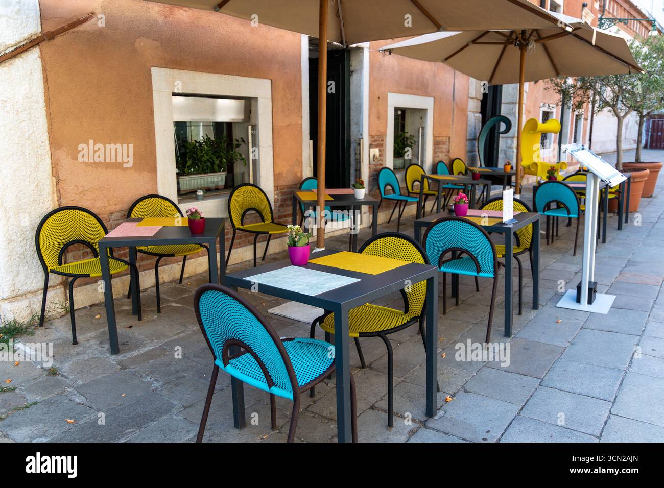 Farbenfrohe Cafés im Freien auf der venezianischen Straße. Helle Stühle und Sonnenschirme schmücken eine moderne Terrasse im Freien entlang einer ruhigen Terrasse Stockfoto