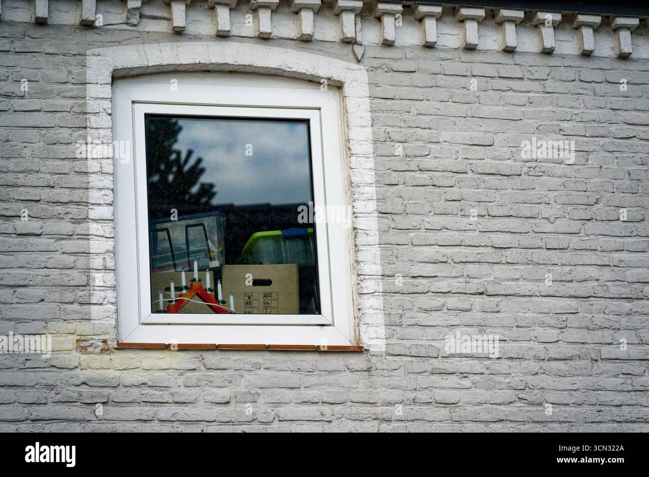 Eine graue Ziegelwand hält ein weiß gerahmtes Kunststofffenster. Im Inneren erhebt sich eine Weihnachtspyramide in dreieckiger Form, während Kästen und Metallkästen gestapelt sind Stockfoto Eine graue Ziegelwand hält ein weiß gerahmtes Kunststofffenster. Im Inneren erhebt sich eine Weihnachtspyramide in dreieckiger Form, während Kästen und Metallkästen gestapelt sind Stockfoto