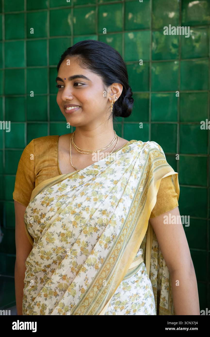 Indische Frau steht und lächelt im Profil vor glänzend grünen Fliesenwänden, die einen floralen Sari tragen Stockfoto