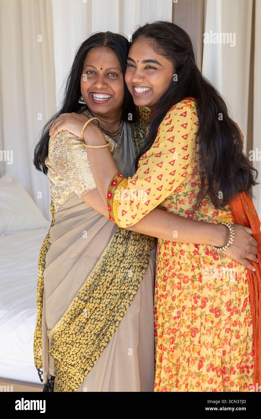 Indische Mutter und Tochter in traditioneller Kleidung umarmen, lächeln im Schlafzimmer mit Schmuck, Bindi Stockfoto