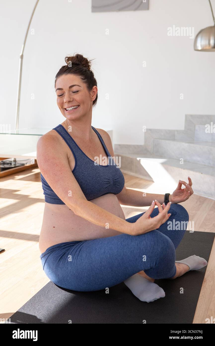 Schwangere Frau sitzend im Kreuz Yoga-Training auf Yogamatte im Wohnzimmer mit Smartwatch Stockfoto