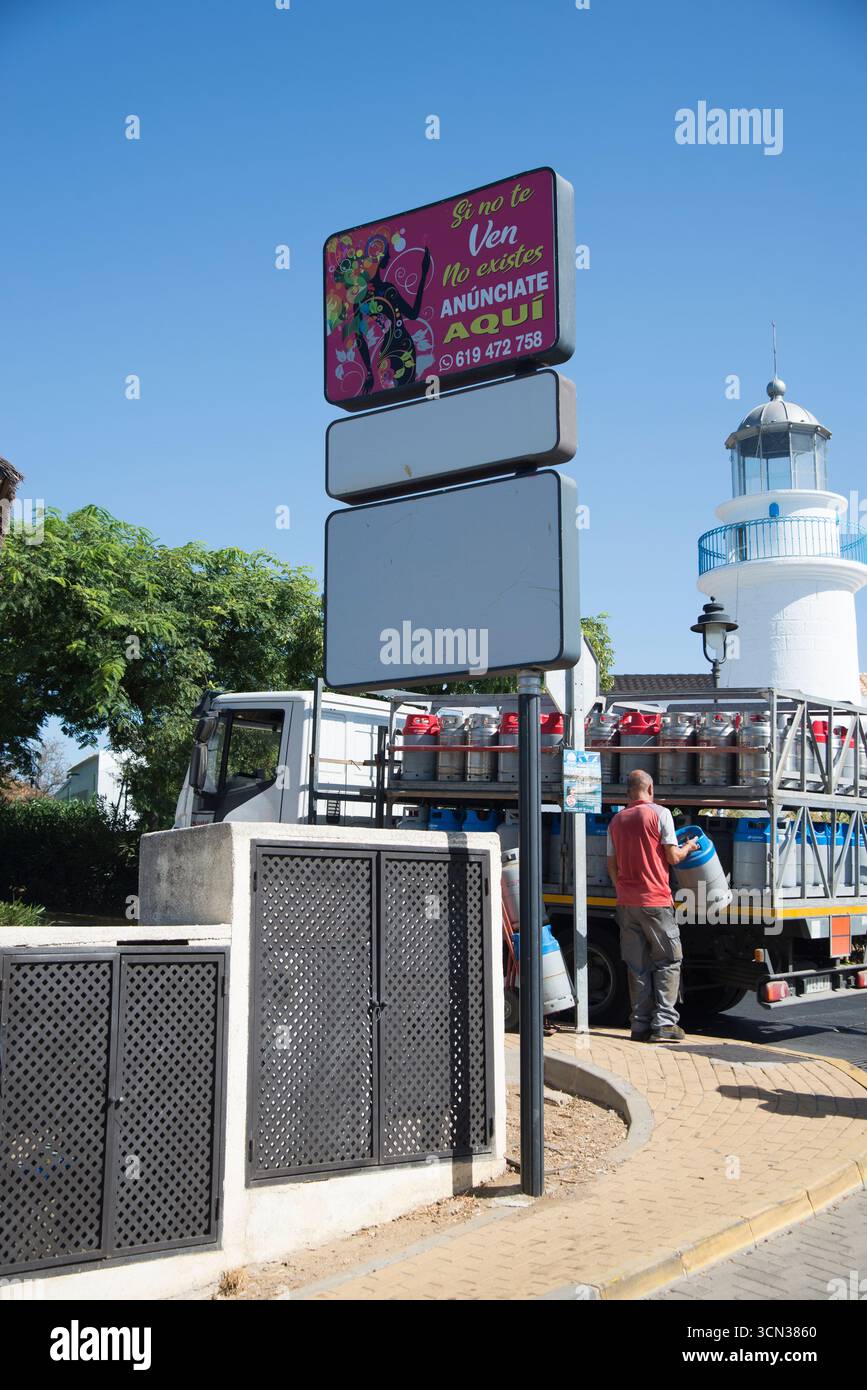 Hombre descargando bombonas de Gas de camión frente al muelle y faro, día soleado, Cartel publicitario, contadores tras puertas en Zona portuaria. Stockfoto