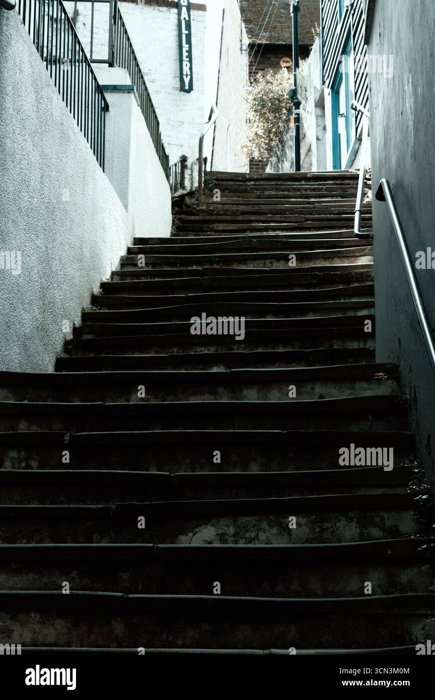 Eine steile, schattige Betontreppe steigt zwischen zwei Mauern in einer urbanen Umgebung nach oben. Die dunklen Stufen deuten auf Herausforderung, Reise und Aufstieg hin. Stockfoto