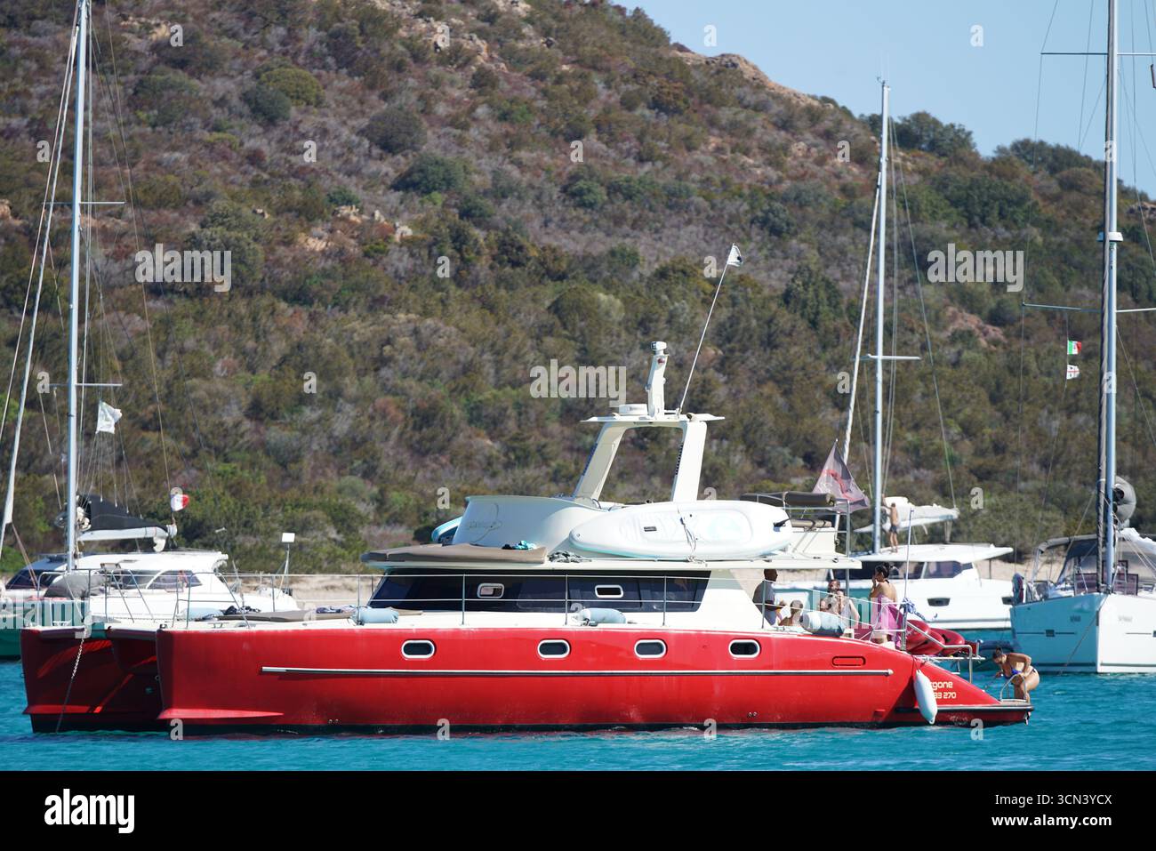 Rote Luxusyacht Vor Anker An Der Sunny Marina Mit Menschen Auf Deck Und Hills Im Hintergrund. Plage de Santa Giulia, Korsika, Frankreich Stockfoto Rote Luxusyacht Vor Anker An Der Sunny Marina Mit Menschen Auf Deck Und Hills Im Hintergrund. Plage de Santa Giulia, Korsika, Frankreich Stockfoto
