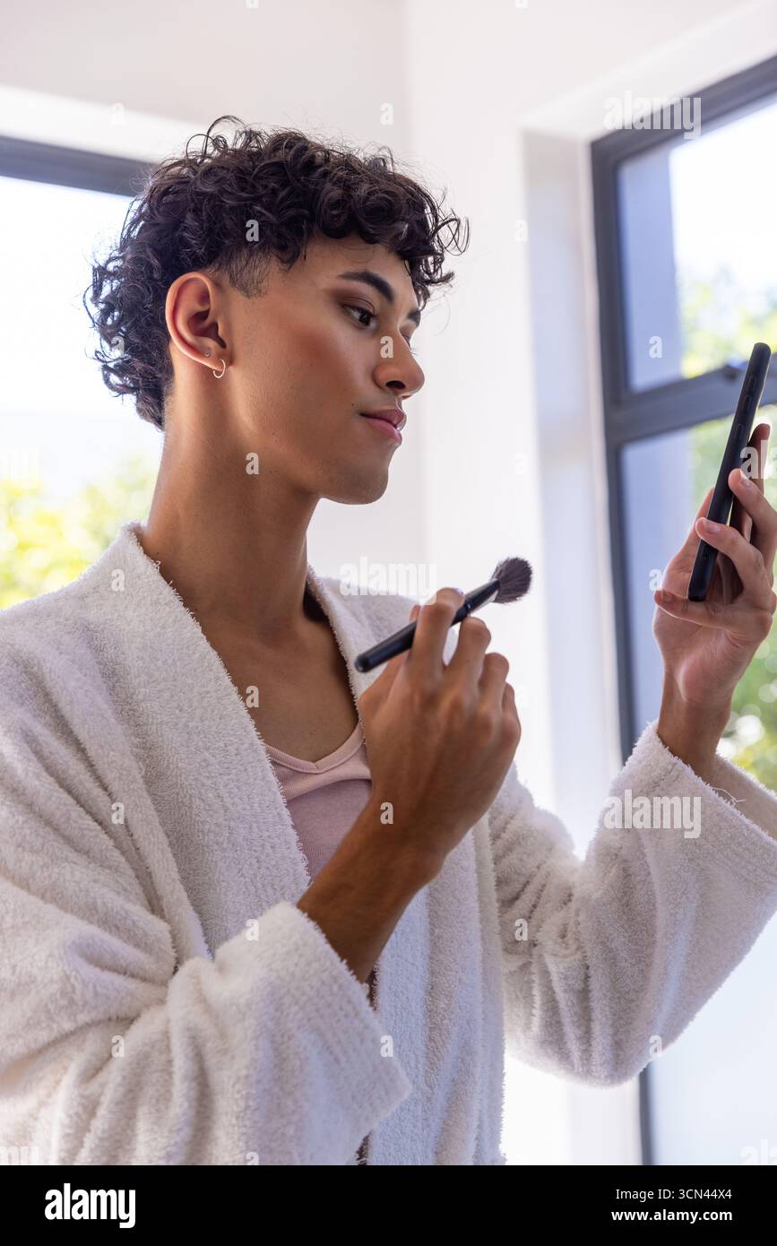 Asiatischer Mann, der Puder mit einem Make-up-Pinsel aufträgt und das Smartphone im Bademantel hält Stockfoto
