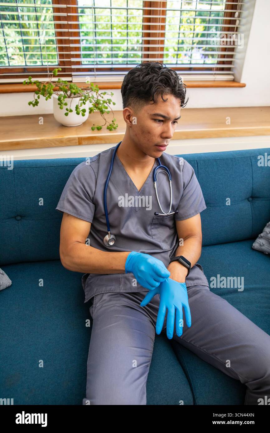 Asiatischer Arbeiter in Peelings mit Stethoskop, der auf Sofa sitzt und Wegwerfhandschuh zieht Stockfoto