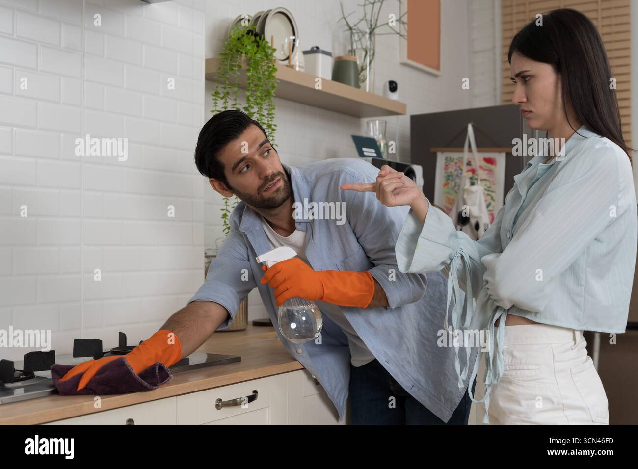 Frustriertes Paar in der Küche, Frau, die mit dem Finger auf den Mann zeigt, der orangefarbene Handschuhe trägt und die Sprühflasche hält, während er den Herd mit einem Tuch reinigt, Konzept von r Stockfoto