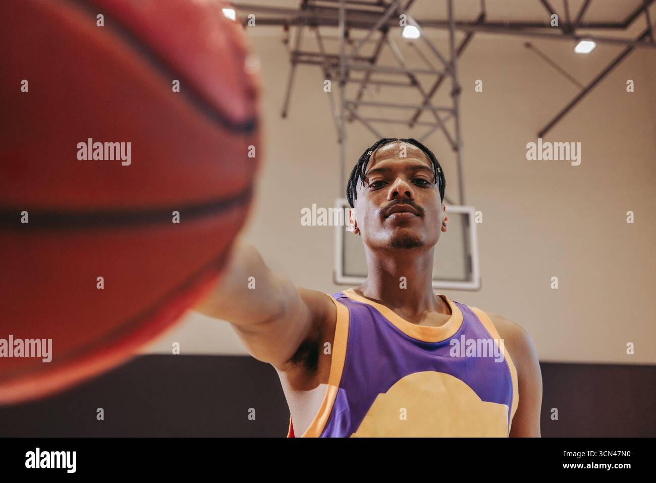 Selbstbewusster junger Mann in Sportbekleidung, der einen Basketball in der Nähe der Kamera in einem Basketballfeld hält und Entschlossenheit, Leichtathletik und konzeptualisiert Stockfoto