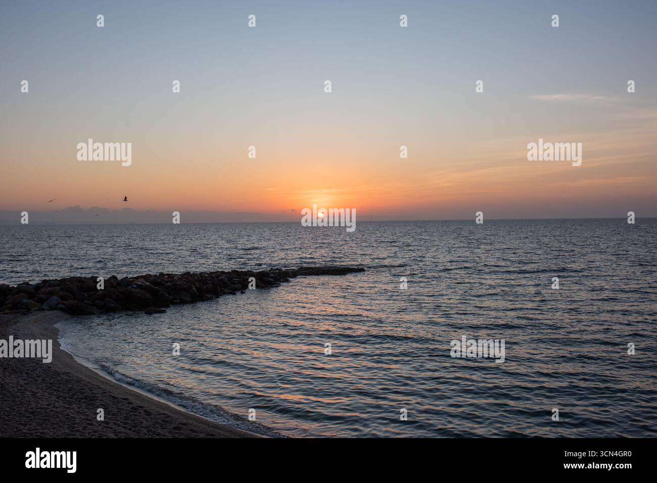 Ein ruhiger Sonnenuntergang über dem Meer mit einem Sandstrand und einem Wellenbrecher. Stockfoto