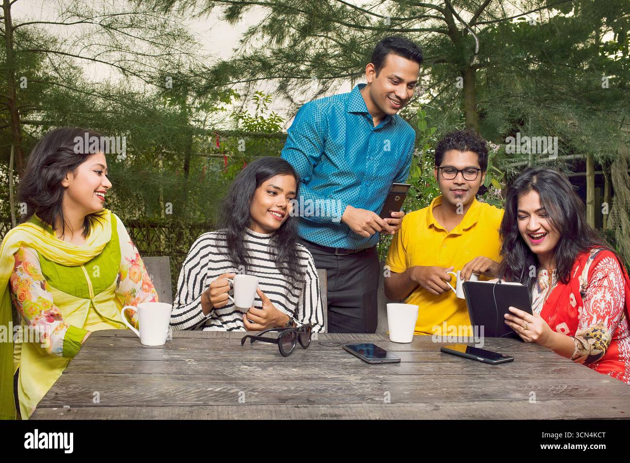 Eine bunte Gruppe von fünf Freunden, eine Mischung aus Männern und Frauen, versammelt sich um einen hölzernen Tisch im Freien. Sie genießen Kaffee und Unterhaltung. Stockfoto