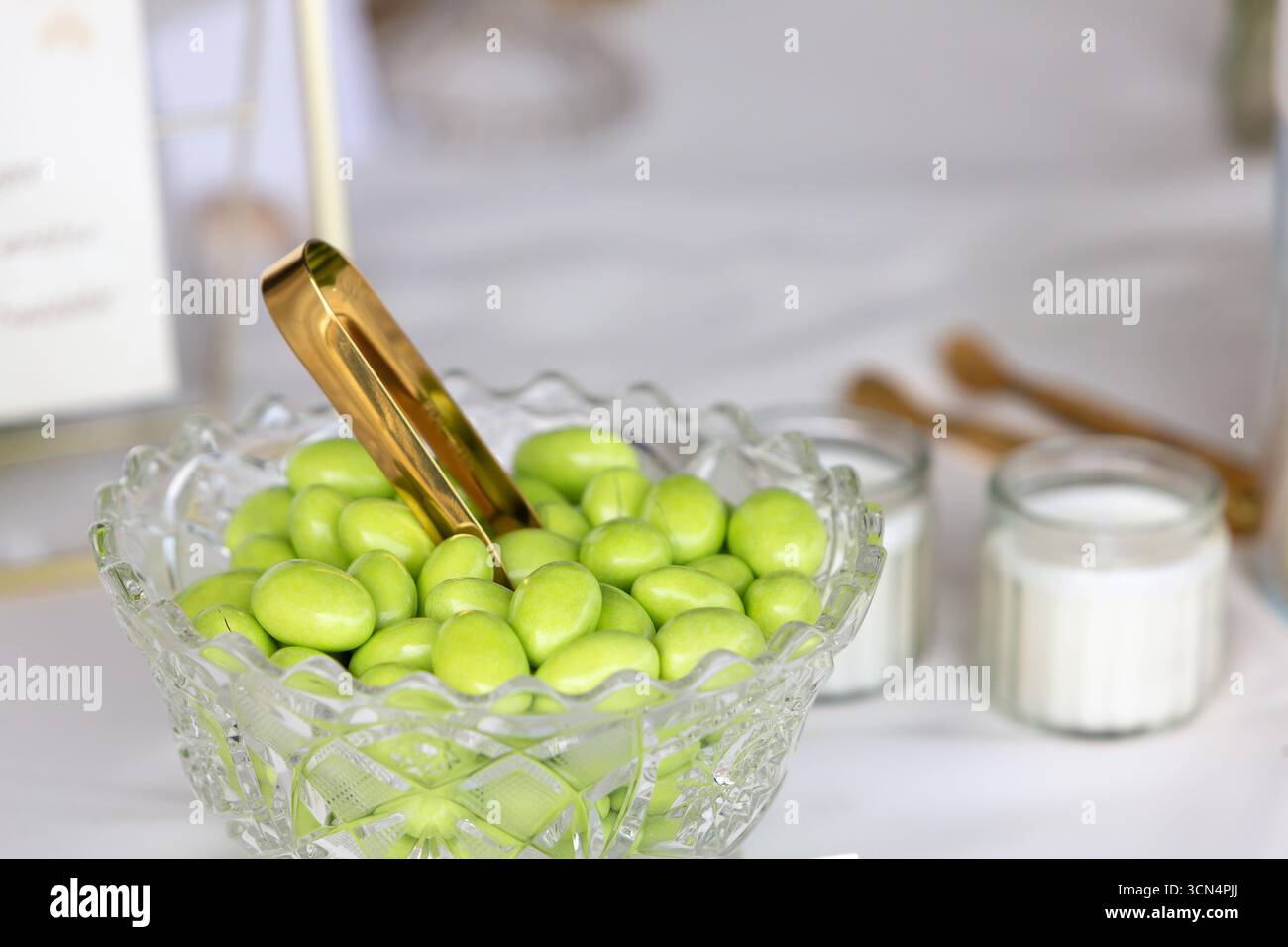 Grüne gezuckerte Mandeln ruhen in einer Kristallschale mit goldener Zange, die Sie bei einer festlichen Feier oder einem Hochzeitsempfang genießen können Stockfoto