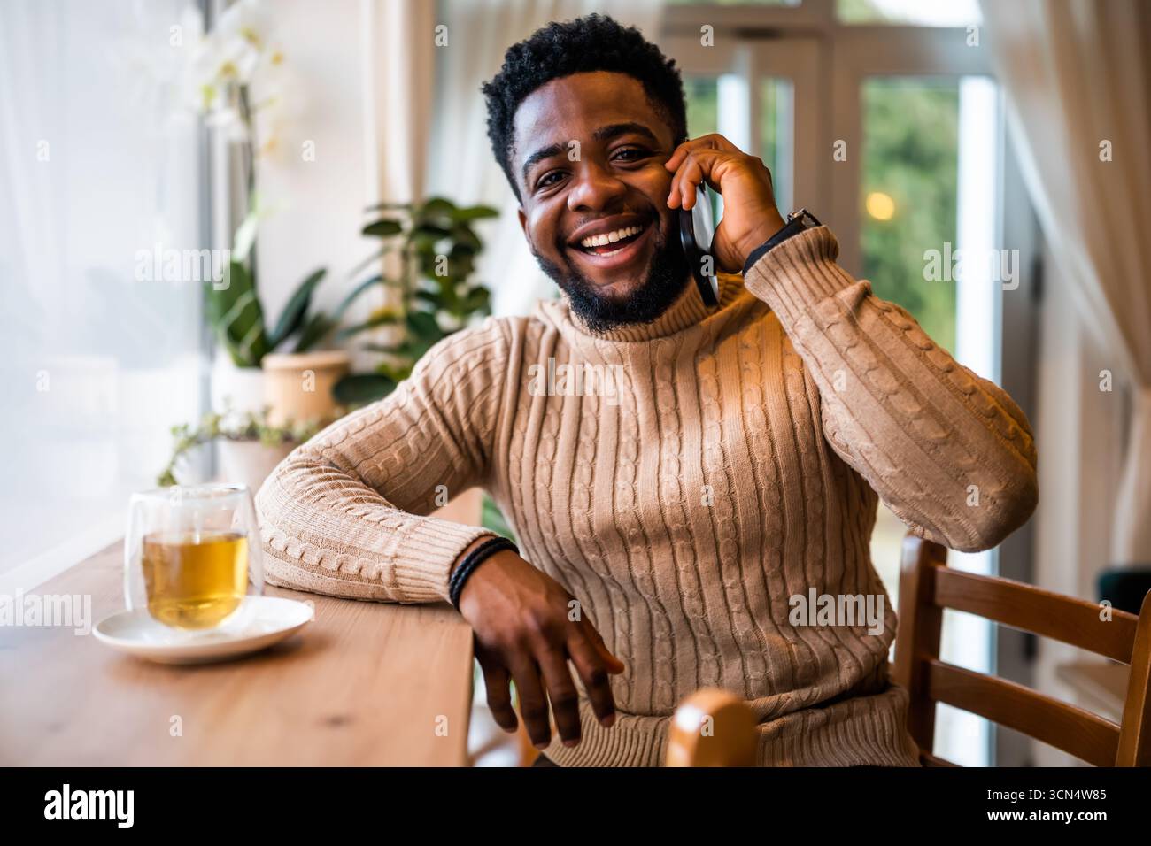 Ein fröhlicher Schwarzer sitzt in lässiger Kleidung an der Bar. Er benutzt das Sprechen auf dem Handy. Stockfoto
