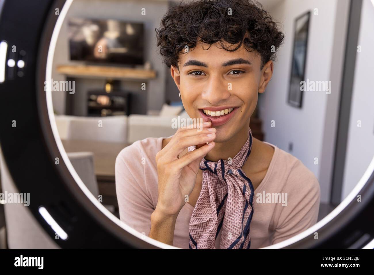 Mann Mitte Zwanziger, der Schal trägt, lächelt in kreisförmiges LED-Ringlicht im Wohnzimmer Stockfoto