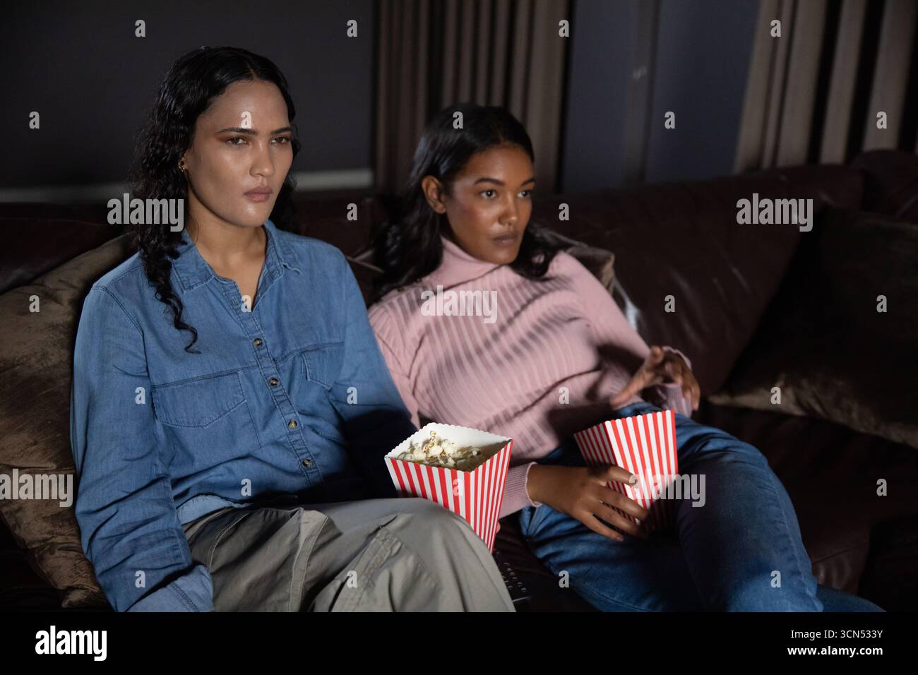Asiatische Freundinnen sitzen auf einem Ledersofa und halten rot-weiße Popcorn-Boxen im Heimkino Stockfoto