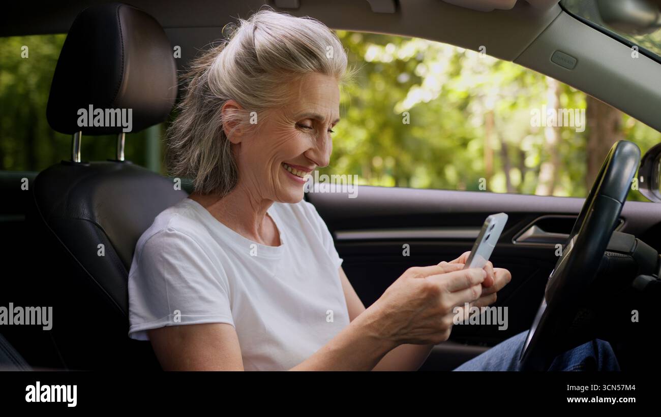 Fröhliche kaukasische reife Frau ältere ältere ältere Frau Rentner Auto Fahrer im Auto mit Handy spielen Spiel Ballen Faust Stockfoto