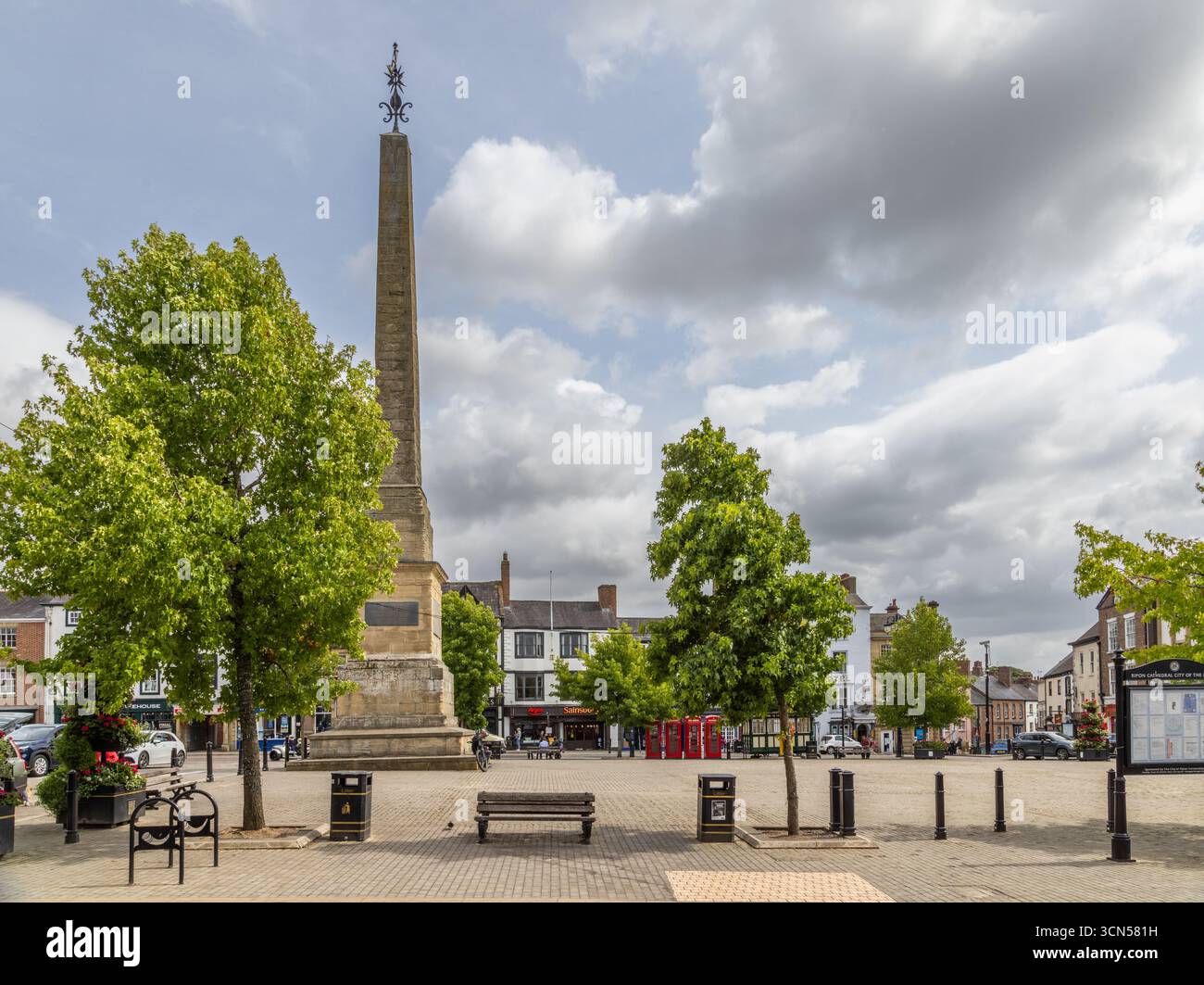 Der Ripon Obelisk auf dem Marktplatz in North Yorkshire, ein denkmalgeschütztes Denkmal aus dem Jahr 1702, umgeben von Bäumen, Bänken, Geschäften und Telefondosen Stockfoto