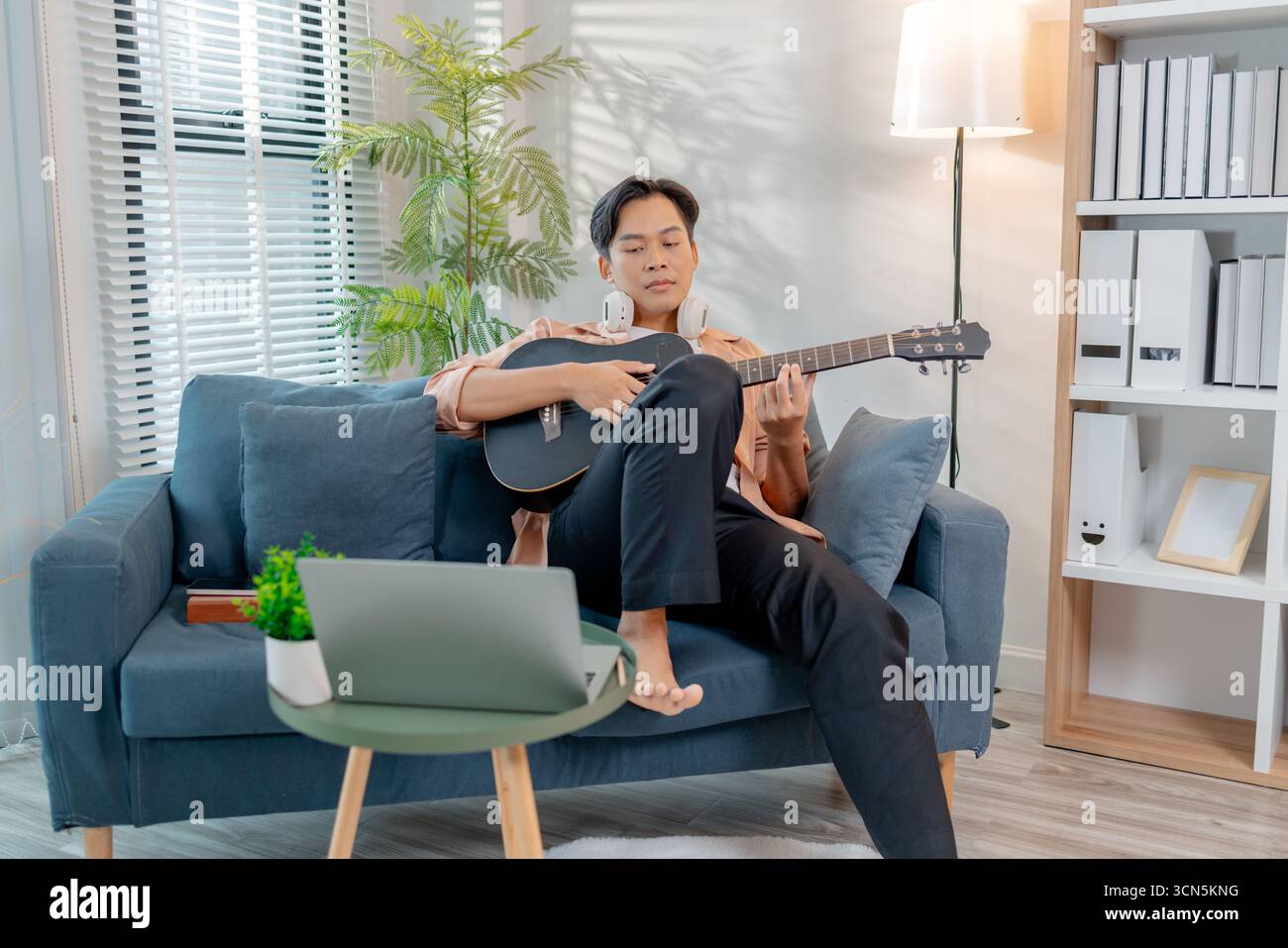 Ein junger Mann spielt fröhlich Gitarre in seinem Wohnzimmer, genießt den Rhythmus, während er Akkorde übt. Seine entspannte Haltung und sein Lächeln spiegeln das Glück wider Stockfoto