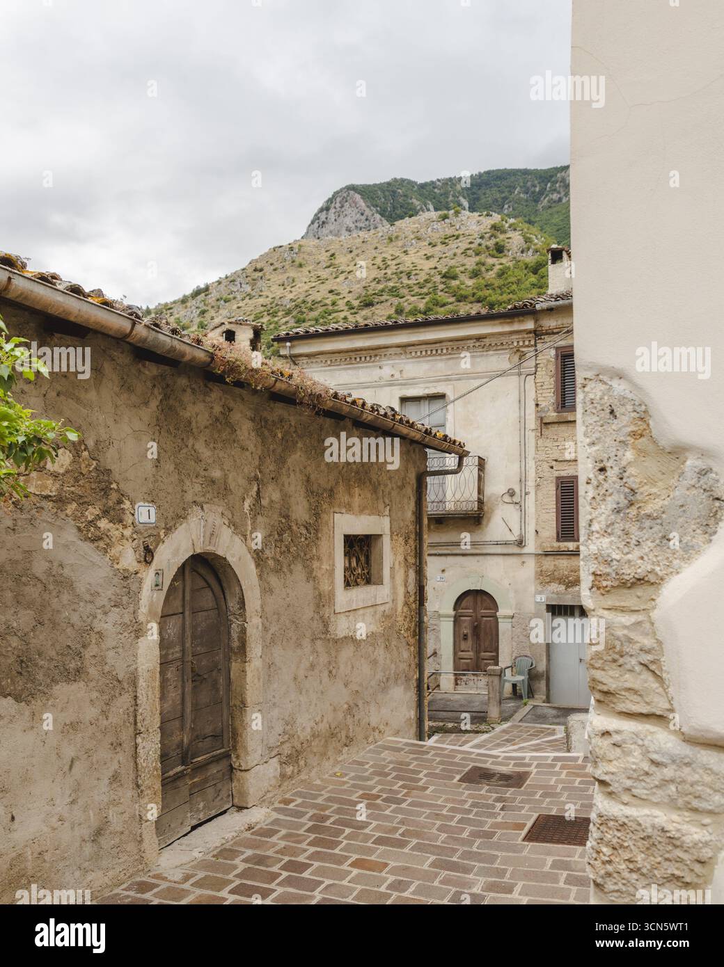 Blick auf eine enge, kopfsteingepflasterte Straße gesäumt von verwitterten, rustikalen Gebäuden, die den Blick auf einen entfernten, felsigen Berg, Anversa degli Abruzzi, Stockfoto