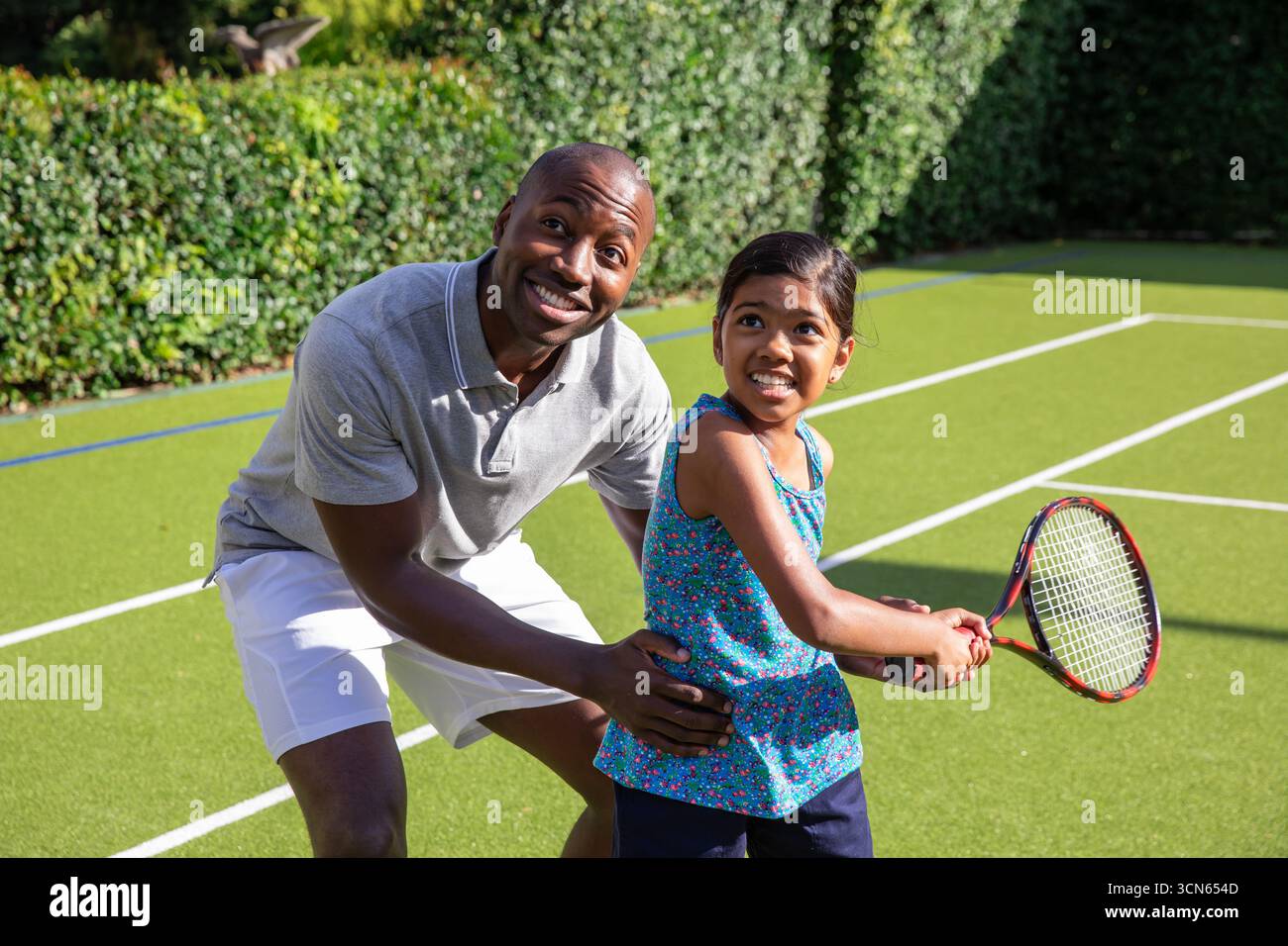 Verschiedene Vater und Tochter knieten beim Tennisschaukeln auf Kunstrasen und halten Schläger Stockfoto