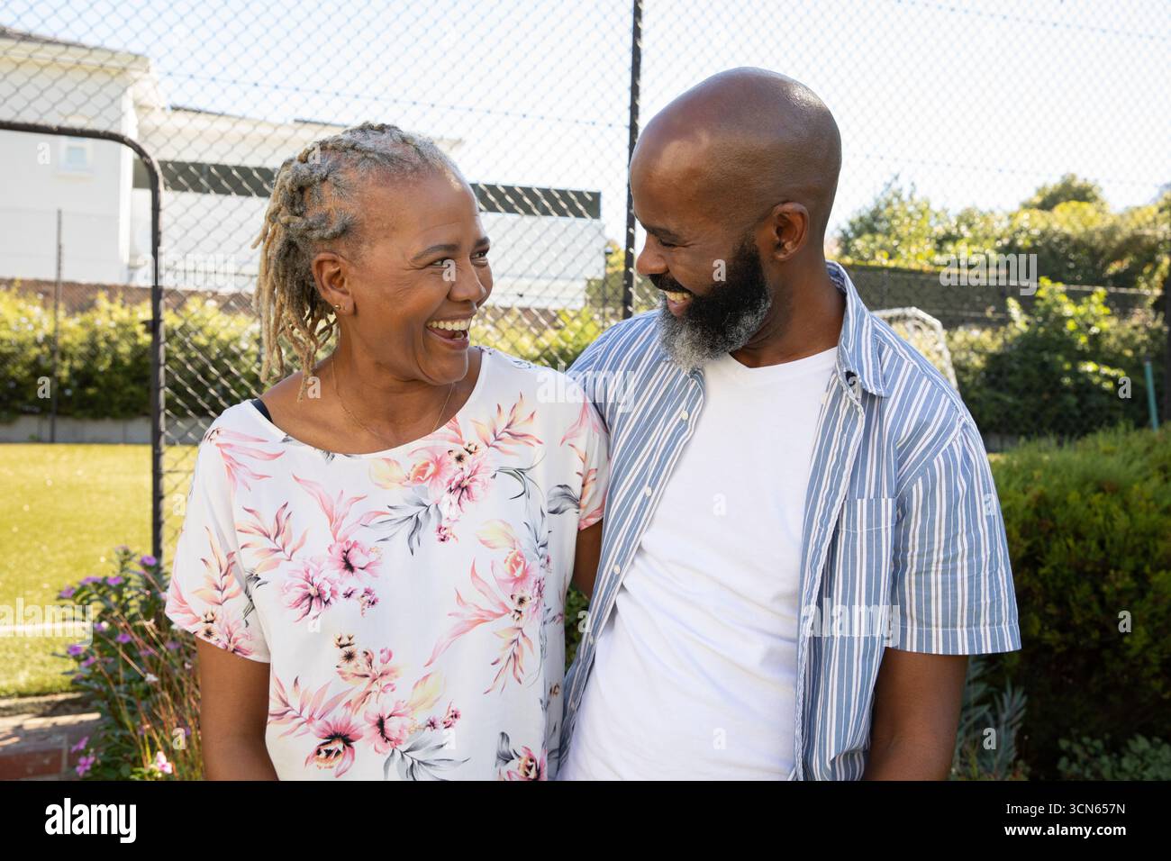 Afroamerikanisches Paar lächelt mit dem Arm um die Schulter, während es am Maschendrahtzaun im Hof steht. Romantisch, Gesellschaft, Natur, glücklich Stockfoto