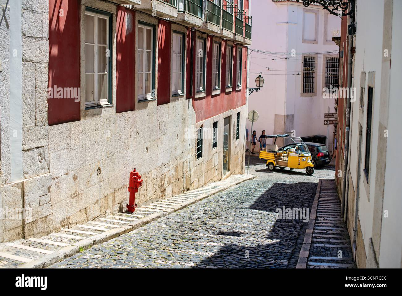 Yellow Tuk Tuk auf der Kopfsteinpflasterstraße in Lissabon Portugal // LISSABON, Portugal — Ein hellgelbes Tuk-Tuk fährt durch eine enge, kopfsteingepflasterte Straße in Lissabon, Portugal. Die Straße ist von traditioneller portugiesischer Architektur gesäumt, mit Gebäuden mit roten und weißen Fassaden und verzierten Balkonen. Auf der linken Straßenseite steht ein roter Hydrant neben einer Steinmauer. Die Szene fängt die charmante und historische Atmosphäre von Lissabons unverwechselbaren Straßen ein. Stockfoto