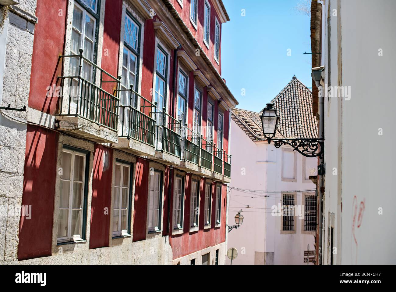 Wohnstraße mit Balkonen Lissabon Portugal // LISSABON, Portugal - Eine Wohnstraße in Lissabon mit traditioneller Architektur mit rot gestrichenen Fassaden und schmiedeeisernen Balkonen. Die Gebäude zeigen charakteristische portugiesische architektonische Elemente, darunter Ziegeldächer und verzierte Fensterrahmen. Diese Aussicht fängt den Charme von Lissabons historischen Vierteln ein, die für ihre engen Gassen und lebhaften Farben bekannt sind. Die Stadt Lissabon ist die Hauptstadt und größte Stadt Portugals und liegt an der Atlantikküste an der Mündung des Flusses Tejo. Stockfoto