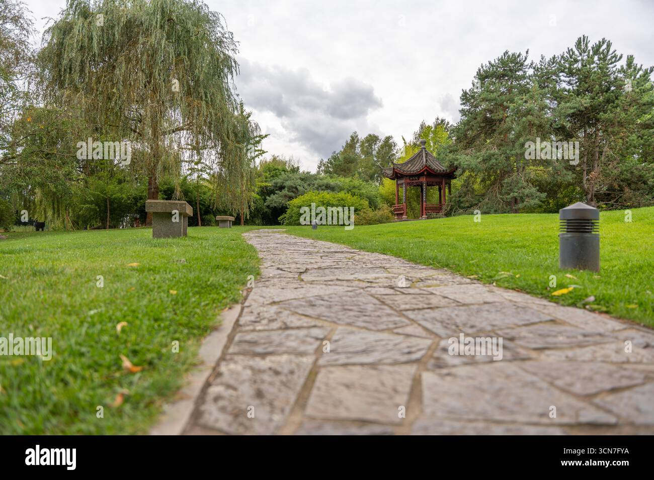 Ein Steinweg führt durch einen Park mit Bänken und einem traditionellen chinesischen Pavillon. Üppige Bäume und Gras umgeben den Gehweg. Stockfoto