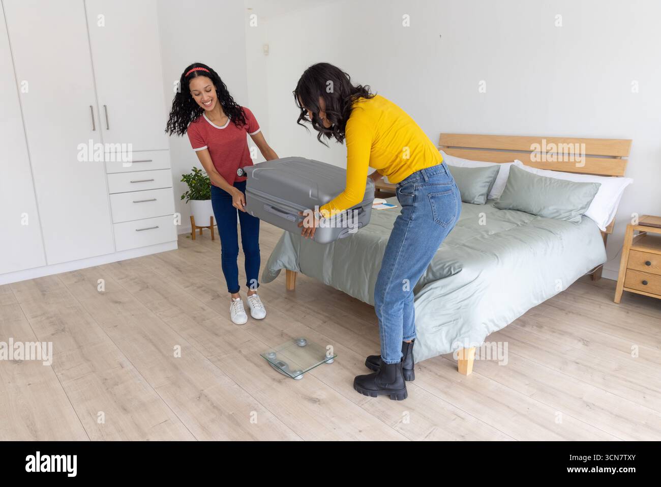 Verschiedene Mutter und Teenager-Tochter legen grauen Koffer auf einer digitalen Waage mit Glasplatte im Schlafzimmer Stockfoto