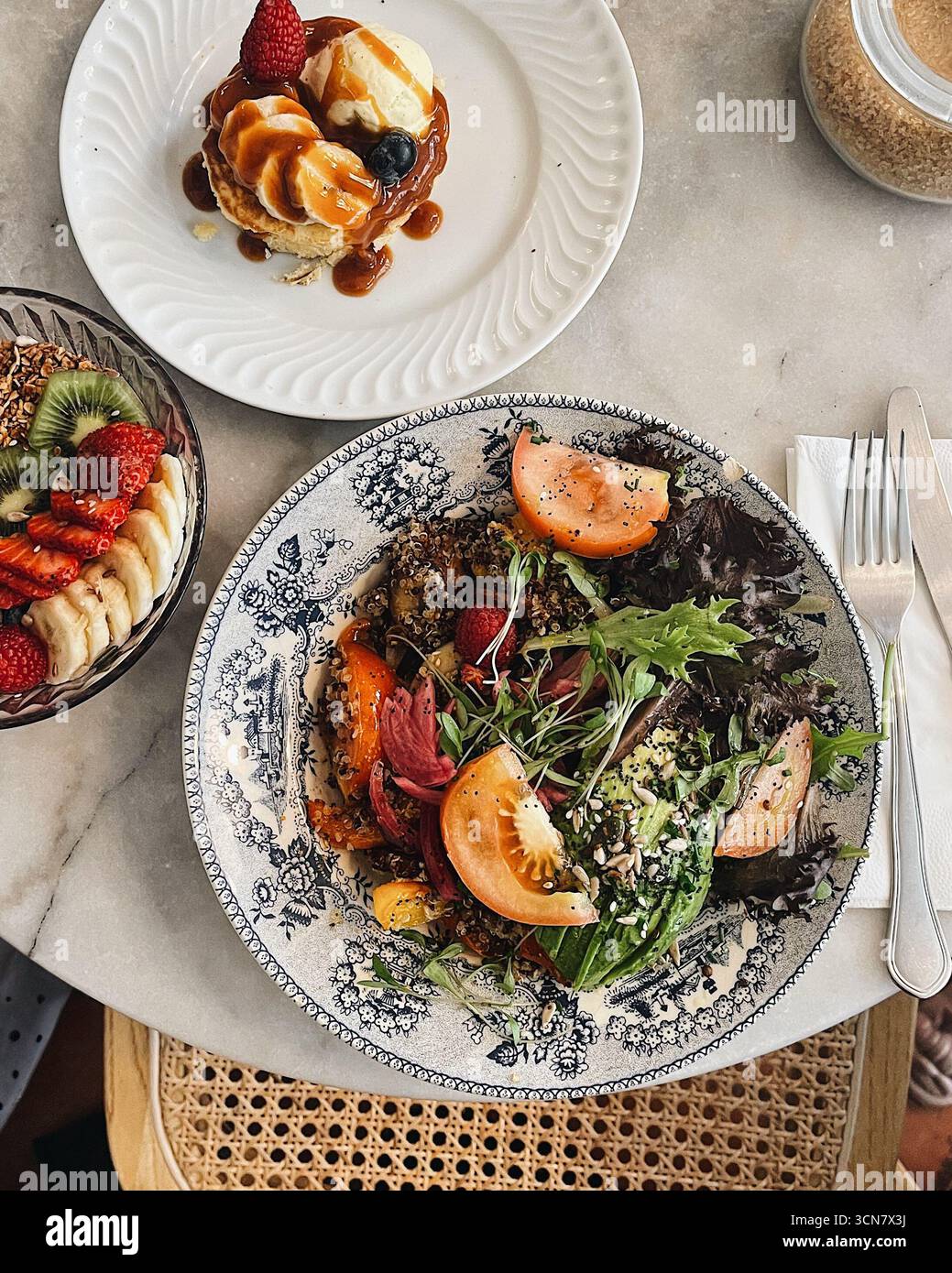 Gourmet Cafe Brunch Aufstrich: Quinoa-Salat, Pfannkuchen mit Karamellsauce und eine gesunde Müsli- und Obstschale mit Kiwi, Erdbeeren und Banane. Stockfoto