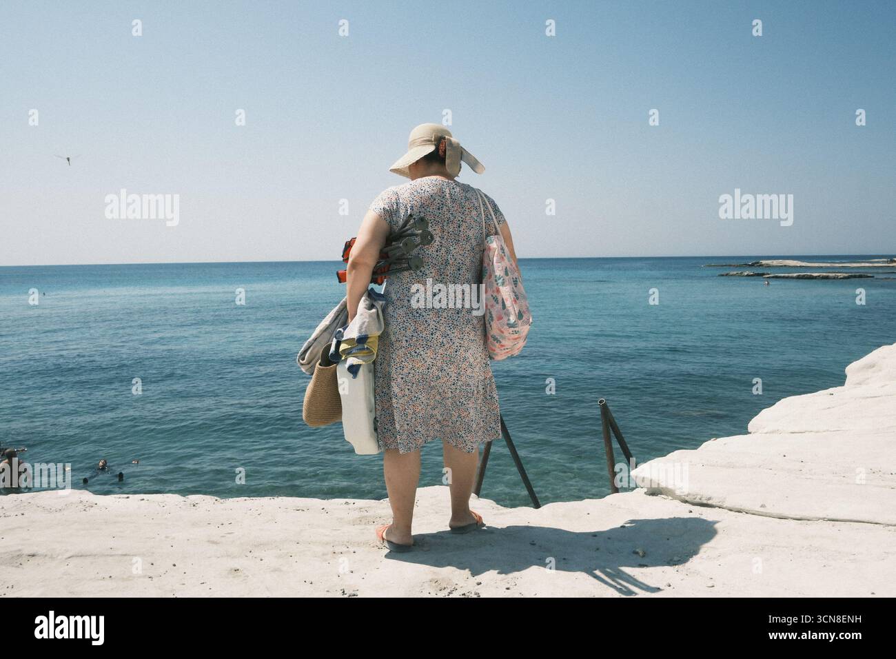 Eine Frau mit Hut, die an einem sonnigen Tag Taschen und Stühle trägt und das Meer beobachtet Stockfoto