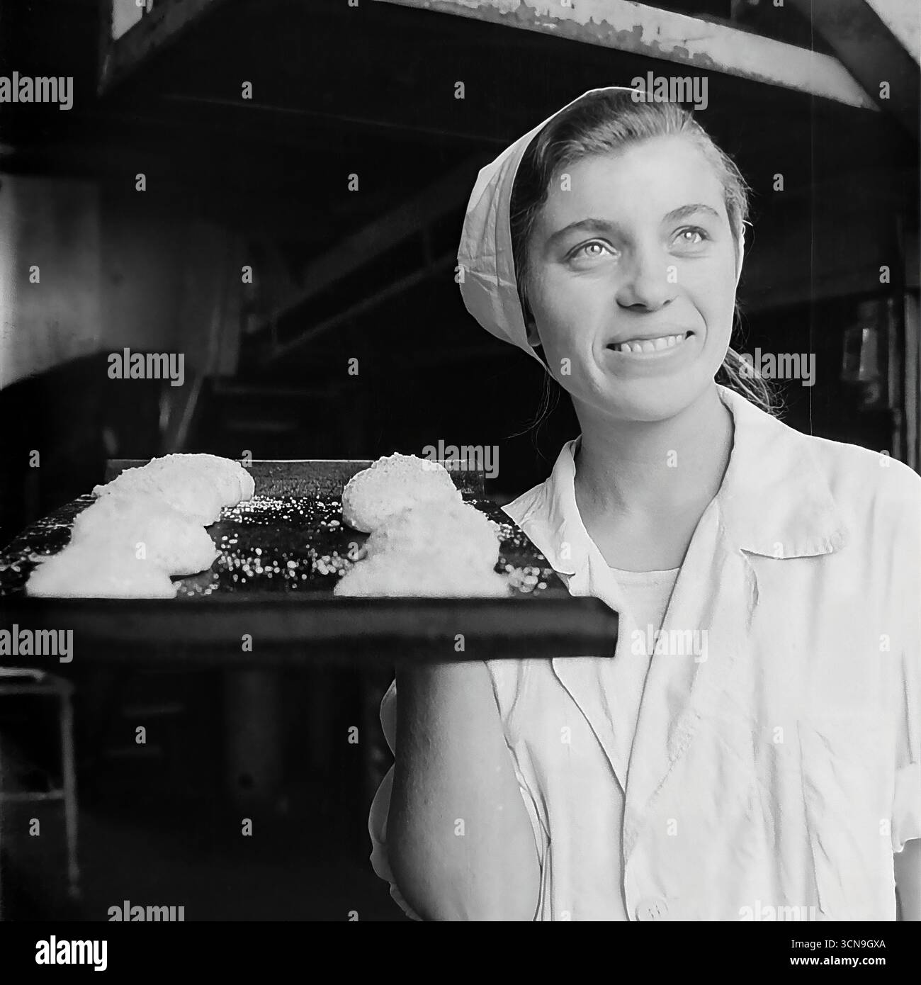 Eine junge, schöne Bäckerin hält ein Backblech mit roher Teig und posiert für ein inspirierendes Porträt in der alten Brotfabrik in Slawjansk, ukrainische SSR, 1970er Ihr hoffnungsvoller Blick, der auf das Licht gerichtet ist, und ihr helles Lächeln schaffen ein idealisiertes Bild einer sowjetischen Arbeiterin, voller Optimismus und Stolz auf ihren Job. Das Foto ist im Stil des sozialistischen Realismus entstanden, der die arbeitende Person glorifiziert Stockfoto