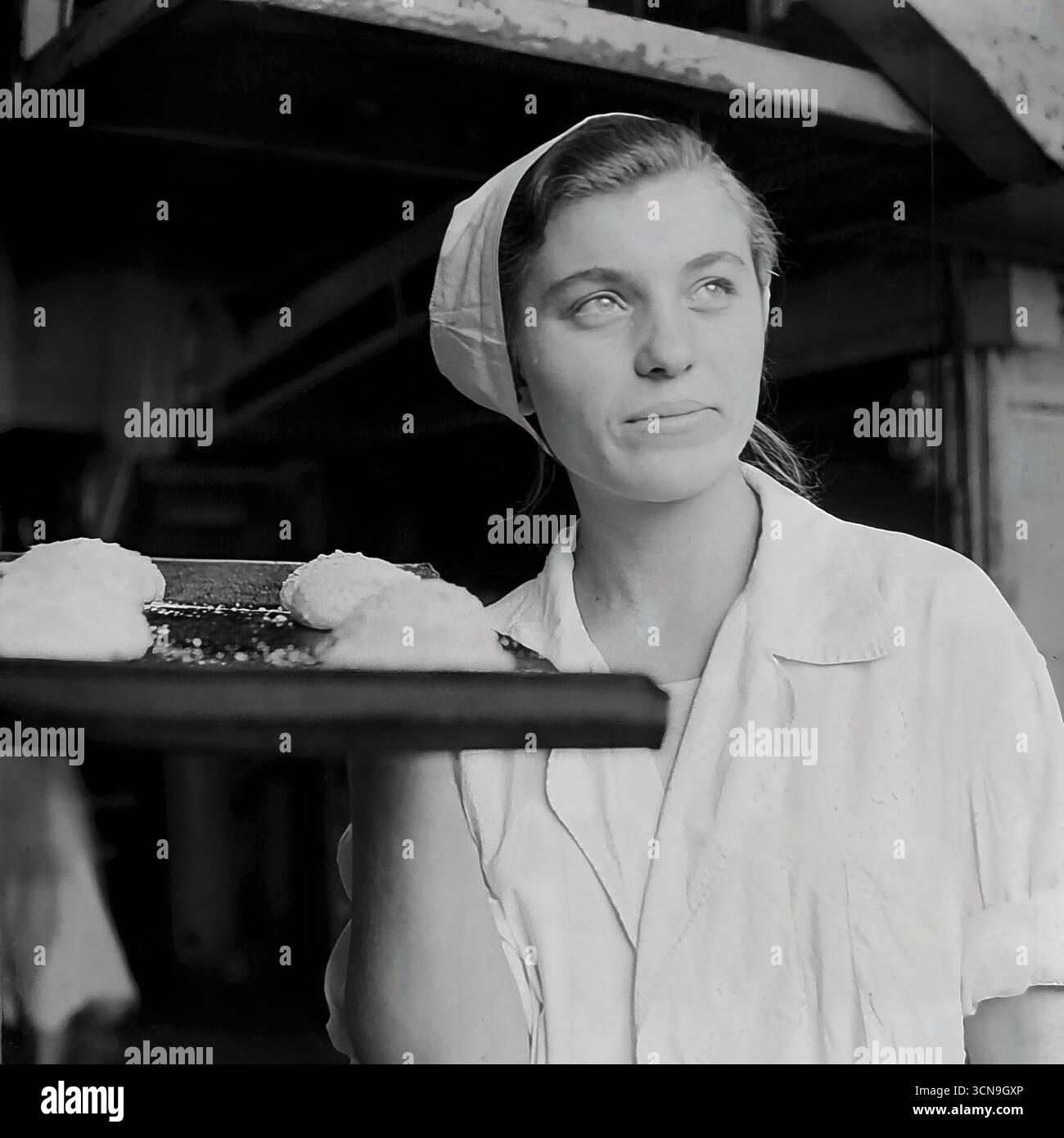 Eine junge, schöne Bäckerin hält ein Backblech mit roher Teig und posiert für ein inspirierendes Porträt in der alten Brotfabrik in Slawjansk, ukrainische SSR, 1970er Ihr hoffnungsvoller Blick, der auf das Licht gerichtet ist, und ihr helles Lächeln schaffen ein idealisiertes Bild einer sowjetischen Arbeiterin, voller Optimismus und Stolz auf ihren Job. Das Foto ist im Stil des sozialistischen Realismus entstanden, der die arbeitende Person glorifiziert Stockfoto