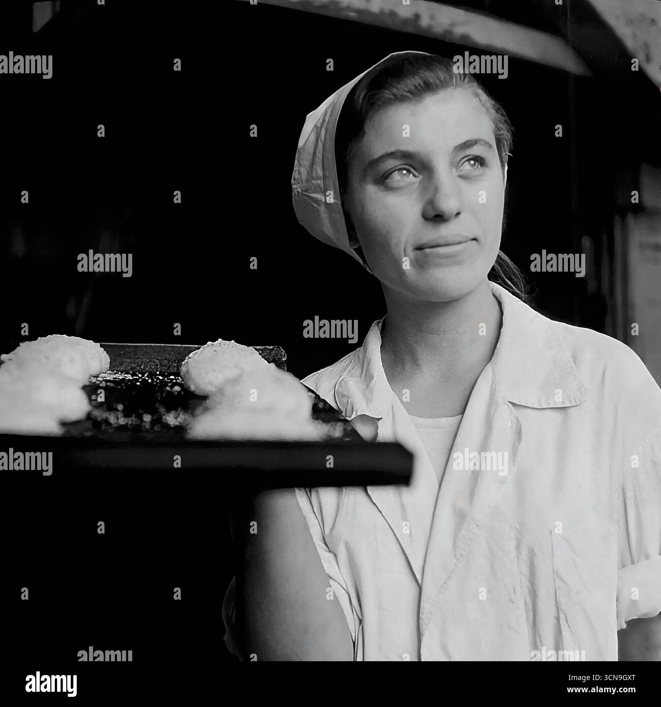 Eine junge, schöne Bäckerin hält ein Backblech mit roher Teig und posiert für ein inspirierendes Porträt in der alten Brotfabrik in Slawjansk, ukrainische SSR, 1970er Ihr hoffnungsvoller Blick, der auf das Licht gerichtet ist, und ihr helles Lächeln schaffen ein idealisiertes Bild einer sowjetischen Arbeiterin, voller Optimismus und Stolz auf ihren Job. Das Foto ist im Stil des sozialistischen Realismus entstanden, der die arbeitende Person glorifiziert Stockfoto