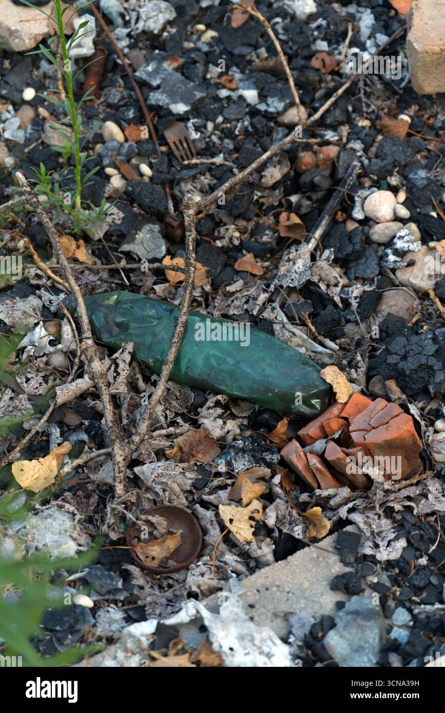 Eine grüne Glasflasche und gebrochenes Terrakotta zwischen Trümmern und verkohlten Überresten auf einem Bett aus trockenen Blättern, Felsen und Schmutz. Stockfoto