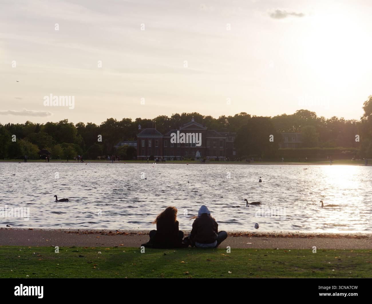 Die Menschen sitzen in den Kensington Gardens, die sich dem Sonnenuntergang nähern, neben einem See mit Schwänen und Kensington Palace dahinter. London. September 2025 Stockfoto