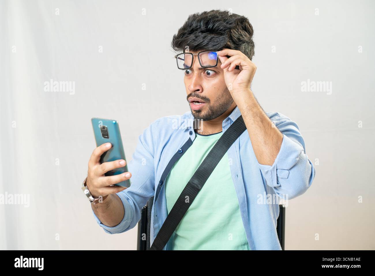 Ein junger Mann mit schockiertem Ausdruck, der seine Brille hält, während er auf sein Handy blickt. Mann überrascht, als er das Telefon ansah und Brillen anstellte. Stockfoto