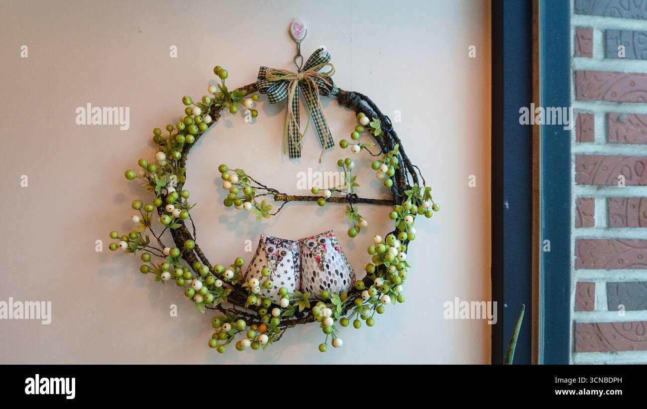 Charmanter rustikaler Kranz mit niedlichen Stoffeulen und grünen Beeren an einer hellen Wand, gemütliche Wohnkultur Stockfoto