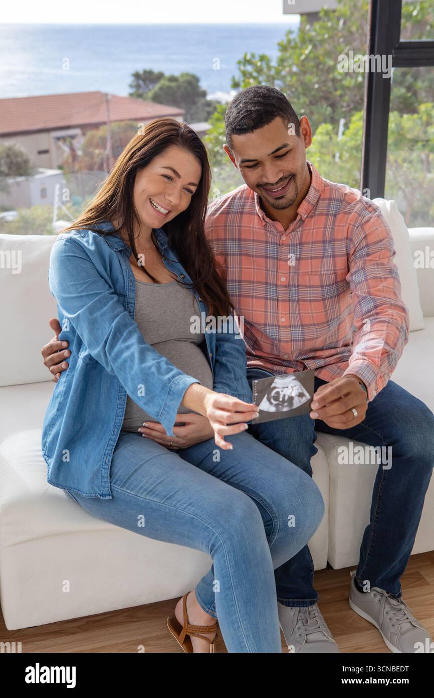 Verschiedene Paare sitzen auf einem weißen Sofa im Wohnzimmer an der Küste und halten einen Ultraschallausdruck am Fenster Stockfoto