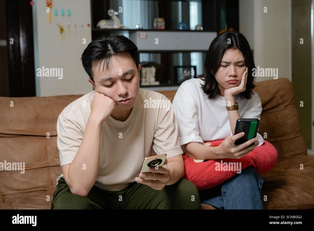 Zwangloses asiatisches Paar, das sich auf einer Couch in einem gemütlichen Wohnzimmer entspannt und auf die Telefone blickt Stockfoto