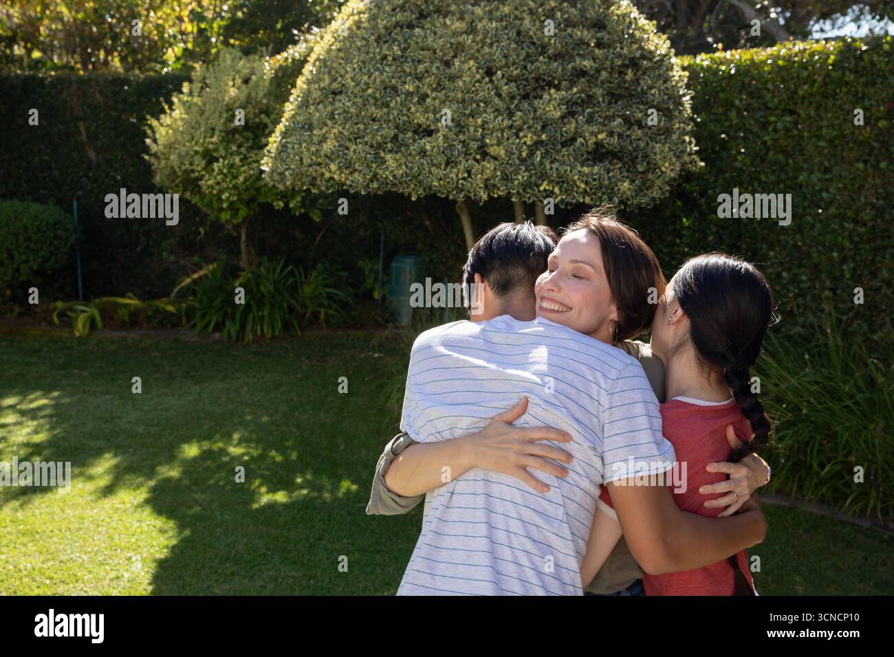 Familie umarmt sich im sonnigen Garten, während das Sonnenlicht durch die getrimmte Hecke gefiltert wird Stockfoto