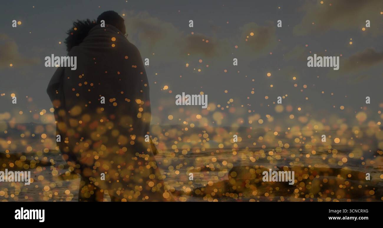 Umarmendes Paar, das bei Sonnenuntergang in eine Decke am Strand gewickelt steht, mit Meereswellen und goldenem Bokeh Stockfoto