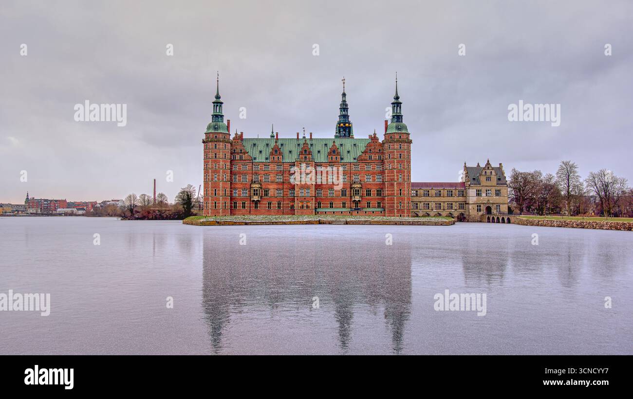 Das majestätische Schloss Frederiksborg spiegelt sich auf einem gefrorenen See unter grauem Winterhimmel und zeigt klassische Renaissance-Architektur im ruhigen Hillerod, den Stockfoto