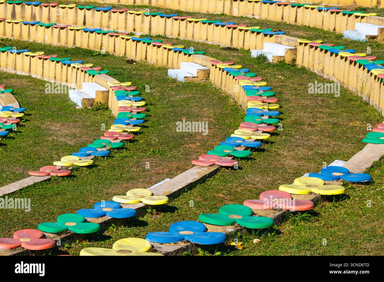 Amphitheater-Sitzgelegenheiten mit hellen, blumenförmigen Hockern aus rotem, blauem, grünem und gelbem Beton, in geschwungenen Reihen im Freien auf grasbewachsenem Boden in d Stockfoto