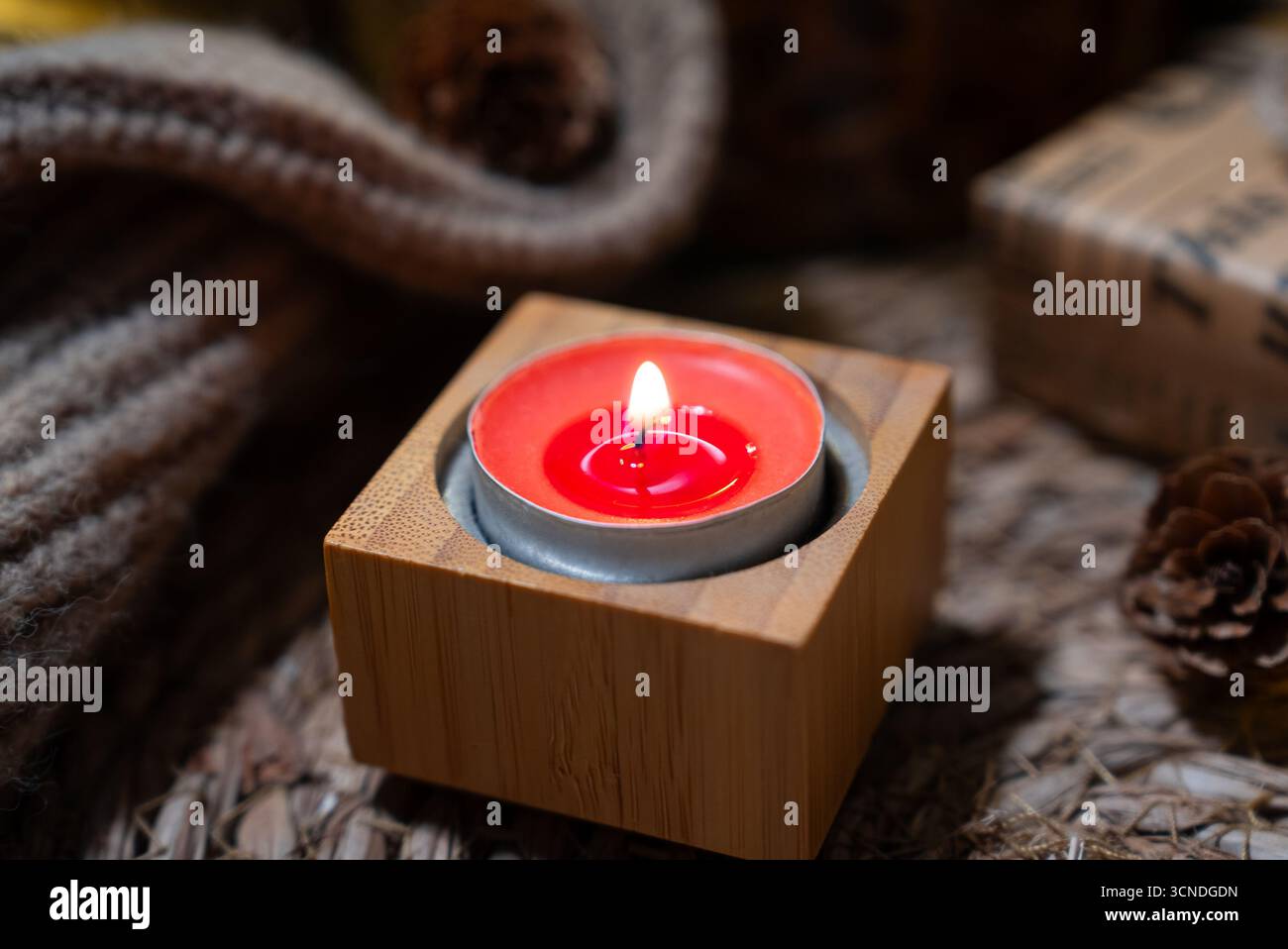 Minimalistische gemütliche Szene mit einer roten Kerze in einem quadratischen Holzhalter auf weicher Strickoberfläche und warmen Bokeh-Lichtern. Stockfoto